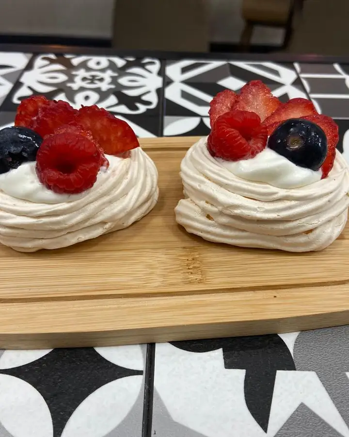 Dos pavlovas con frutos rojos sobre una tabla, en una mesa de baldosas blancas y negras